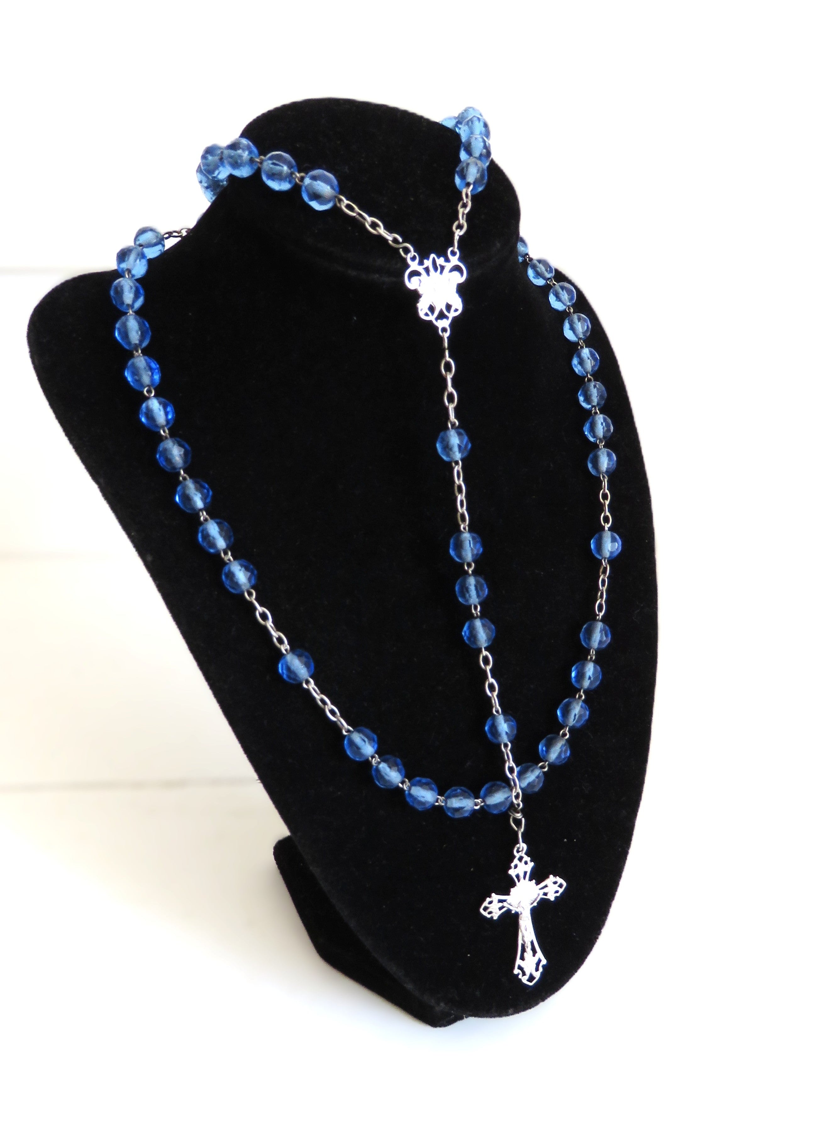 Blue beaded rosary with silver cross on a black stand against a white background