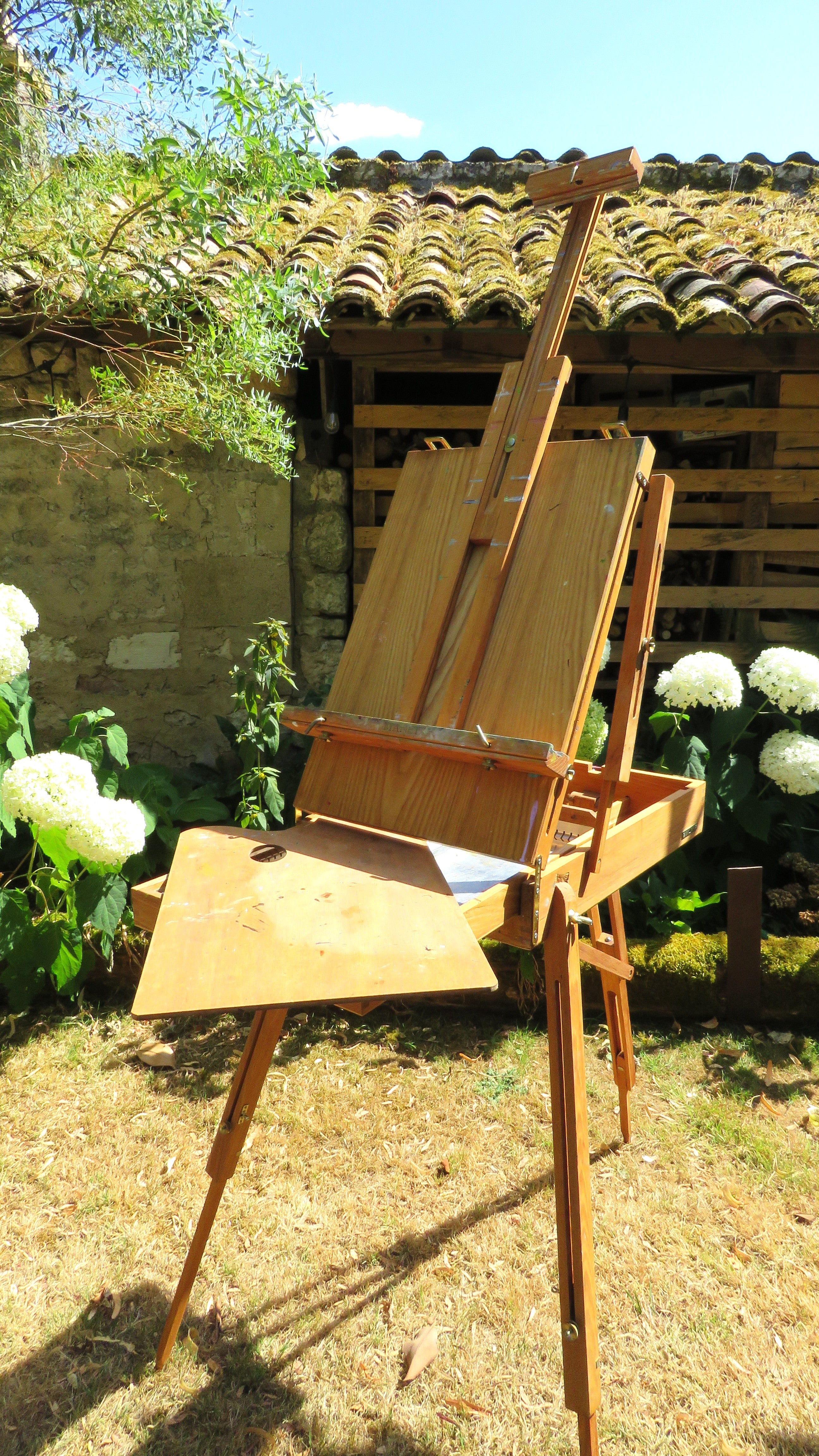 Wooden artist's easel in a garden setting with flowers and a rustic building.