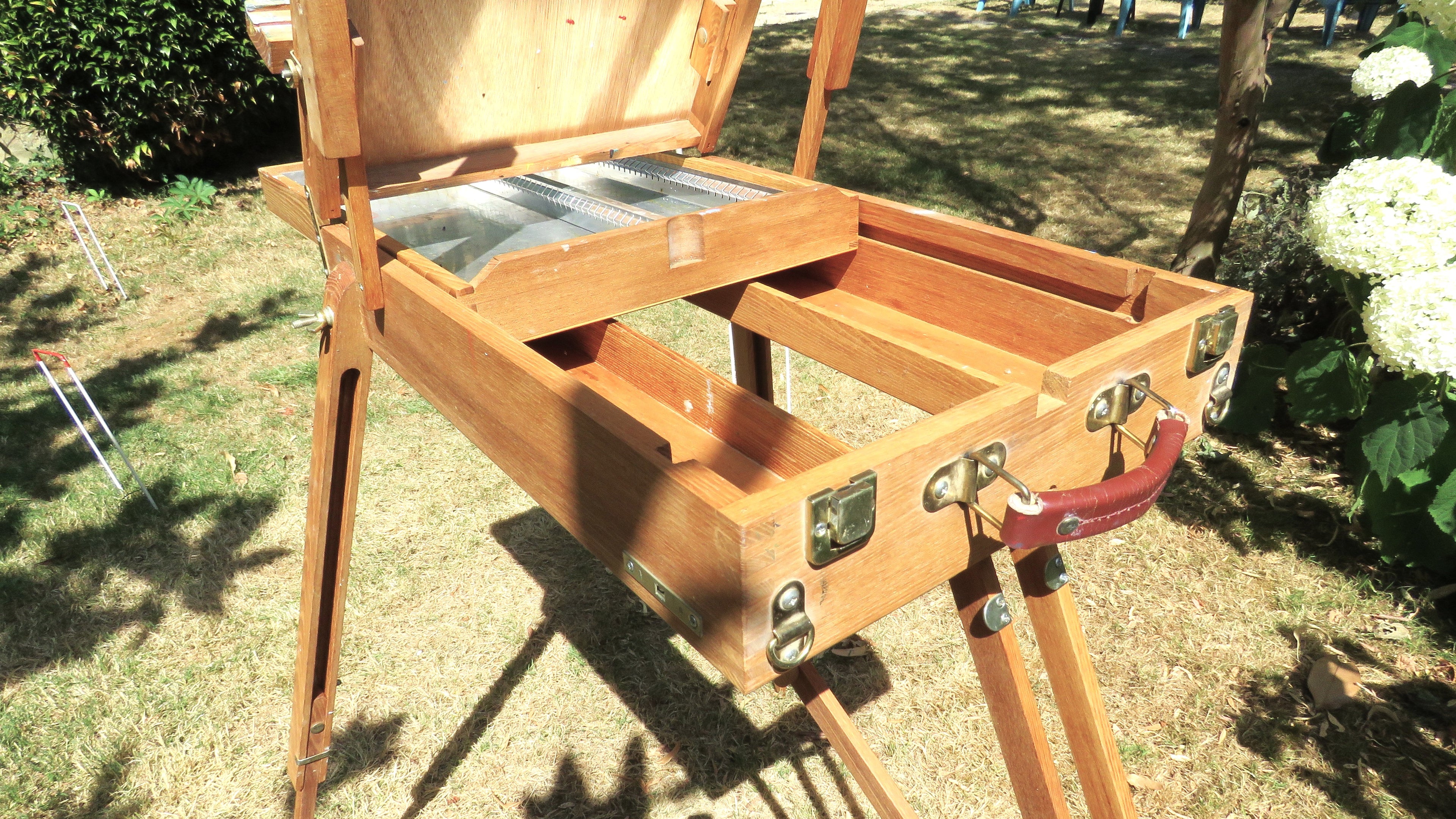 Wooden artist's easel in a garden setting with flowers and a rustic building.