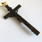 A dark metal crucifix shown on a plain white background with a partial view of a cream-colored beaded rosary in the upper left corner.