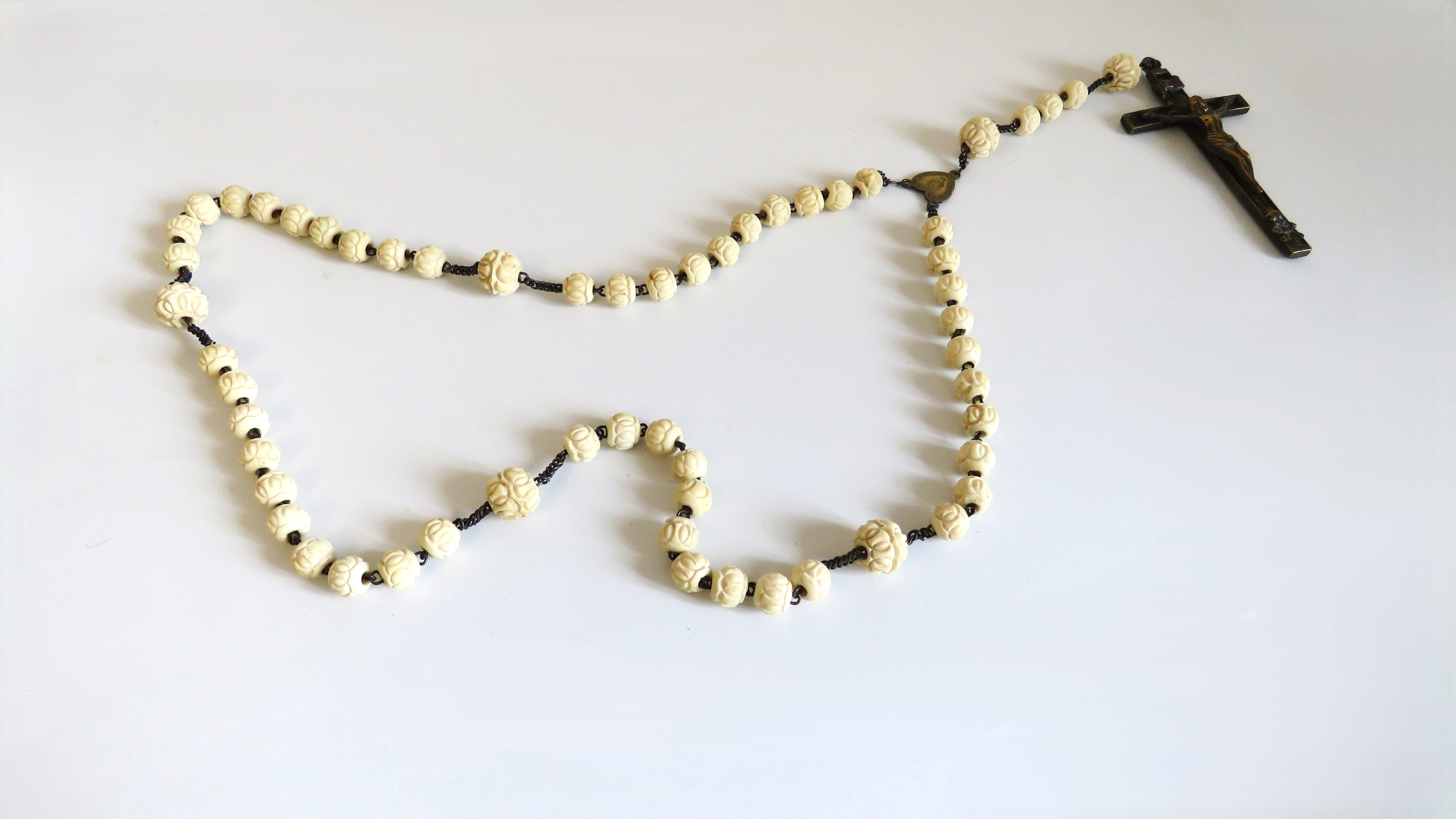 A rosary with cream-colored bone beads featuring a dark metal crucifix and centerpiece, laid out on a plain white surface.