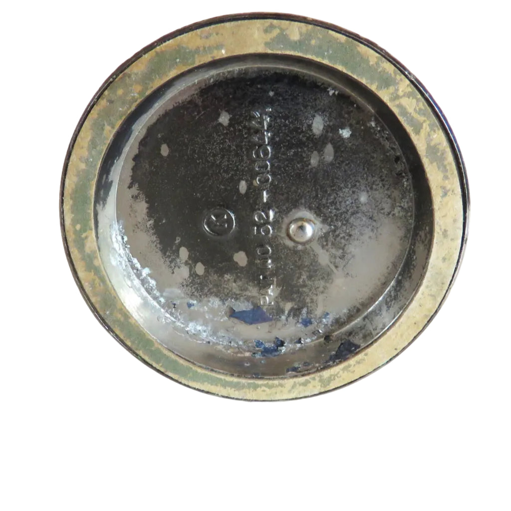A round vintage metal object, possibly a decorative plate or lid, shown from above on a white background. The item features a dark, distressed interior with visible wear, scuffs, and embossed text, surrounded by a weathered gold-toned rim.