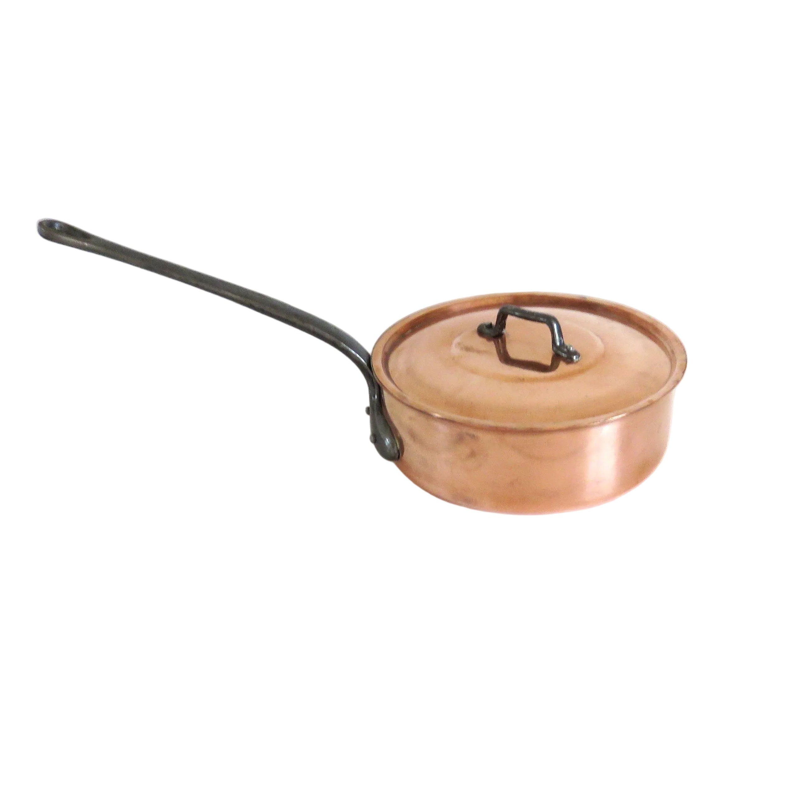 A copper saucepan with a matching lid and a long black handle, resting on a white wooden table against a white wall.