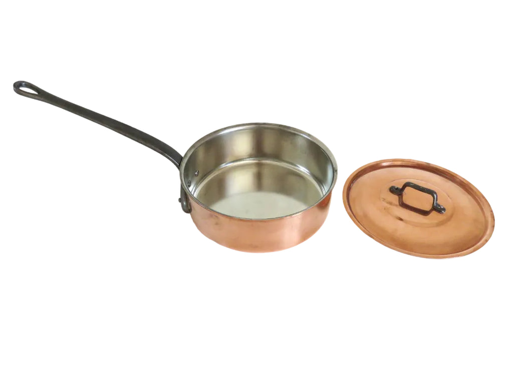 A copper and stainless steel saucepan with a black handle sits next to its matching copper lid on a white wooden table against a white wall.