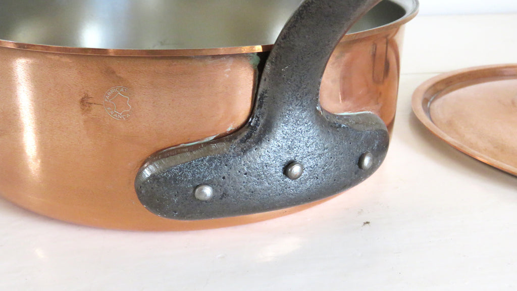 Close-up of a copper saucepan with a dark cast iron handle and rivets, sitting on a white surface with a copper lid partially visible on the right.