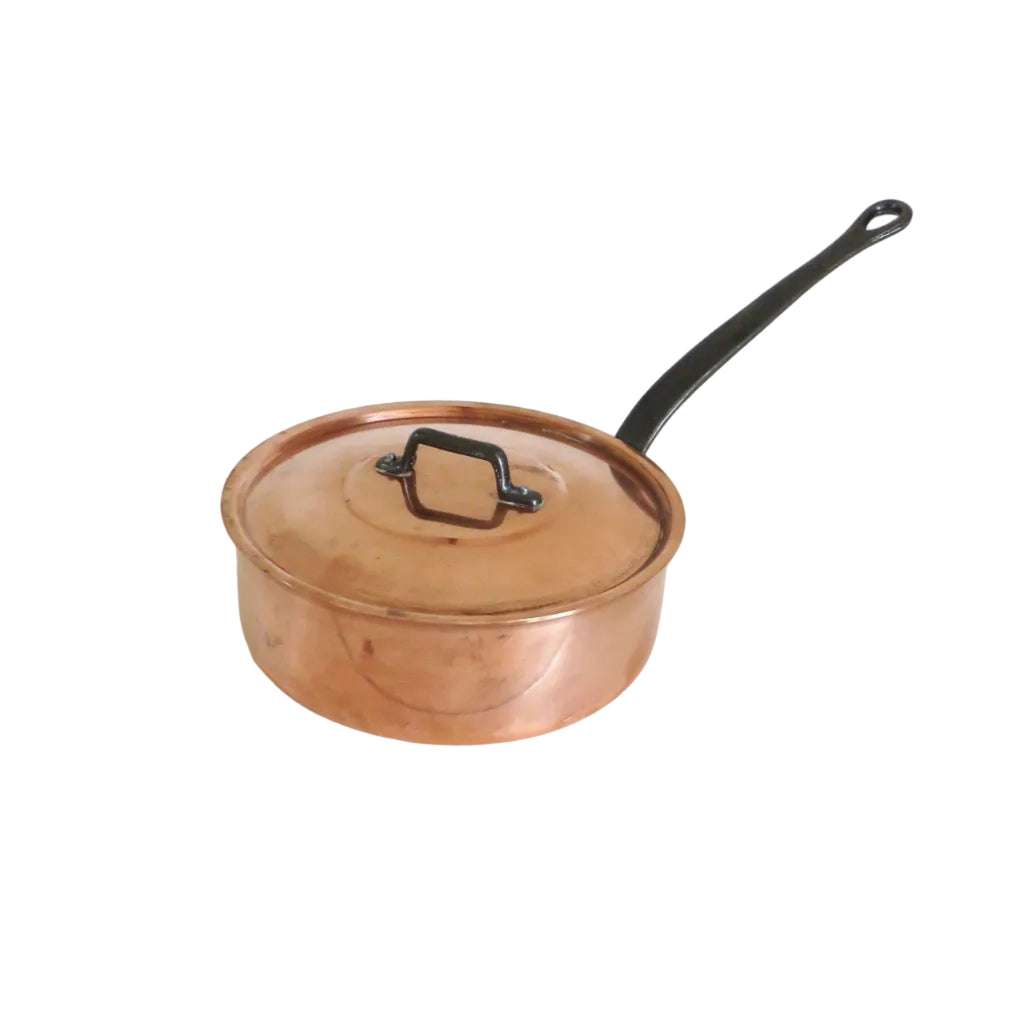 A copper saucepan with a matching lid and a long black handle, resting on a white wooden surface against a white wall.