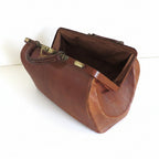 A vintage brown leather doctors gladstone bag with brass hardware, shown open on a white wooden surface against a white background.