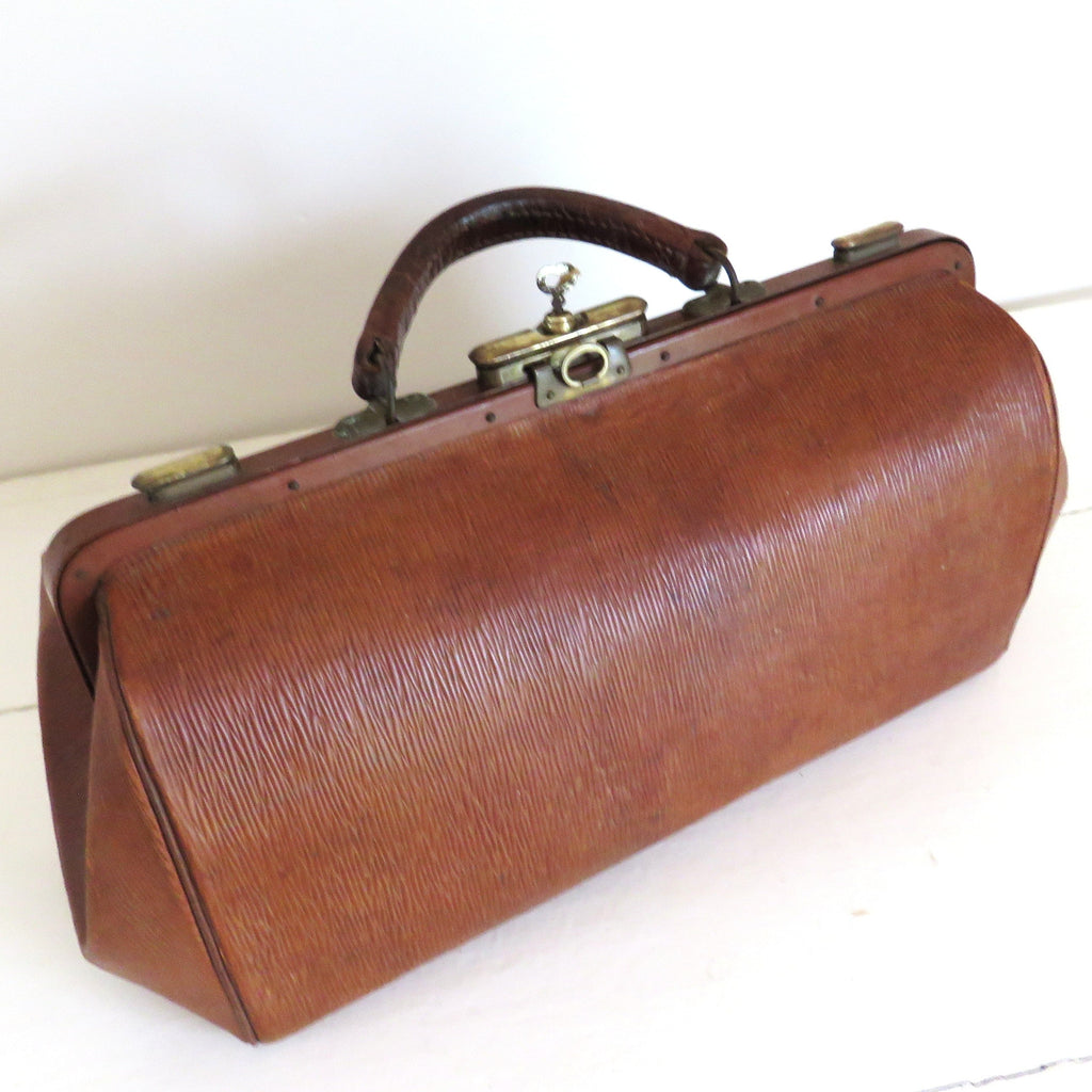 A vintage brown leather doctors bag with a top handle and brass clasp, resting on a white tiled surface.
