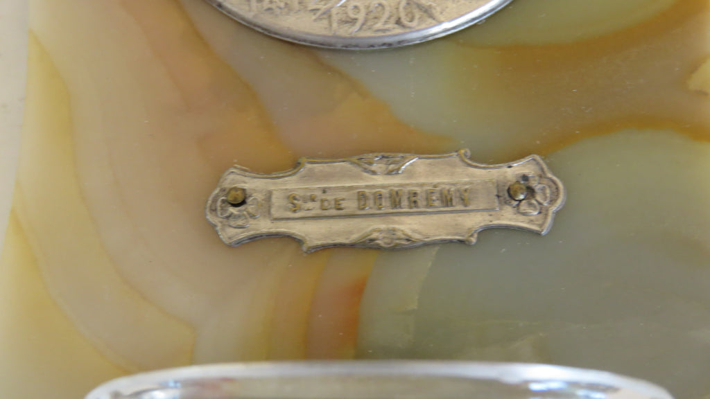 Close-up of a vintage metal brand plaque engraved with 'S. de DOMREMY' mounted on a marbled cream and amber stone surface.