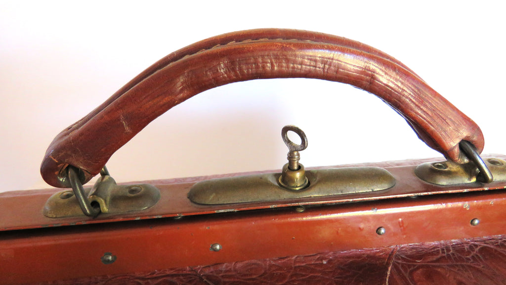 Close-up of a vintage reddish-brown leather suitcase handle with brass hardware and a lock, set against a plain light background.