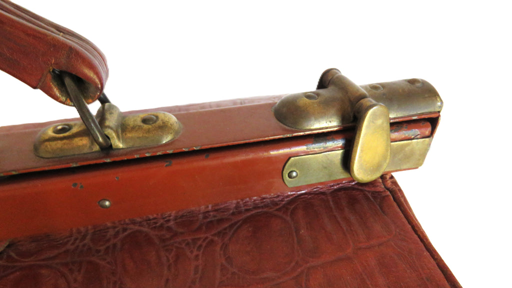 Close-up of a vintage reddish-brown leather briefcase or suitcase corner with brass latches and a textured crocodile-embossed surface, photographed against a white background.