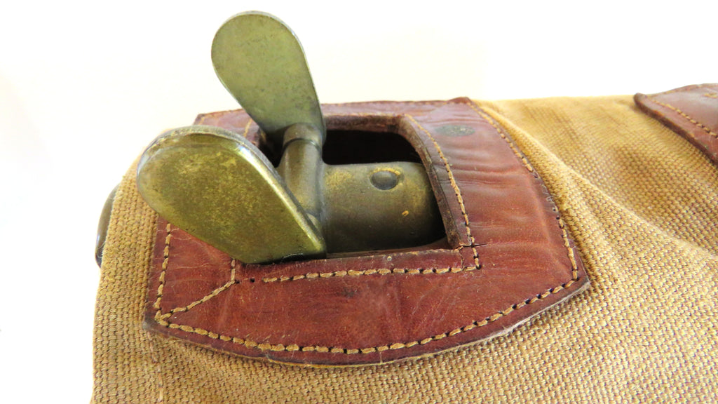 Close-up of a vintage canvas and leather bag with brass hardware, showing a tan woven fabric body, brown leather trim, and a brass loop or clasp on a white background.