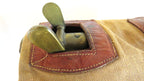 Close-up of a vintage canvas and leather bag with brass hardware, showing a tan woven fabric body, brown leather trim, and a brass loop or clasp on a white background.