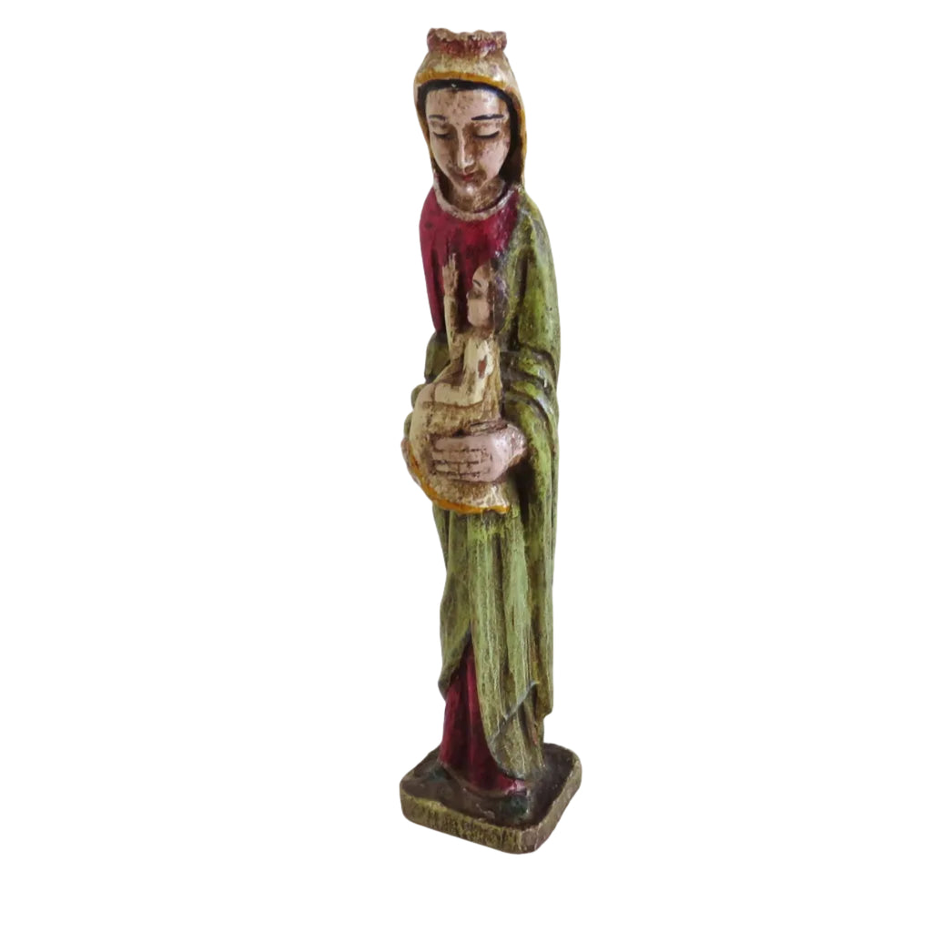 A small carved religious figurine of the Virgin Maryrobed holding a child, standing on a plain white surface against a white wall.