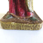 Close-up of the base and lower robe of a figurine, showing a textured gold base with a red and green garment, on a plain white background.