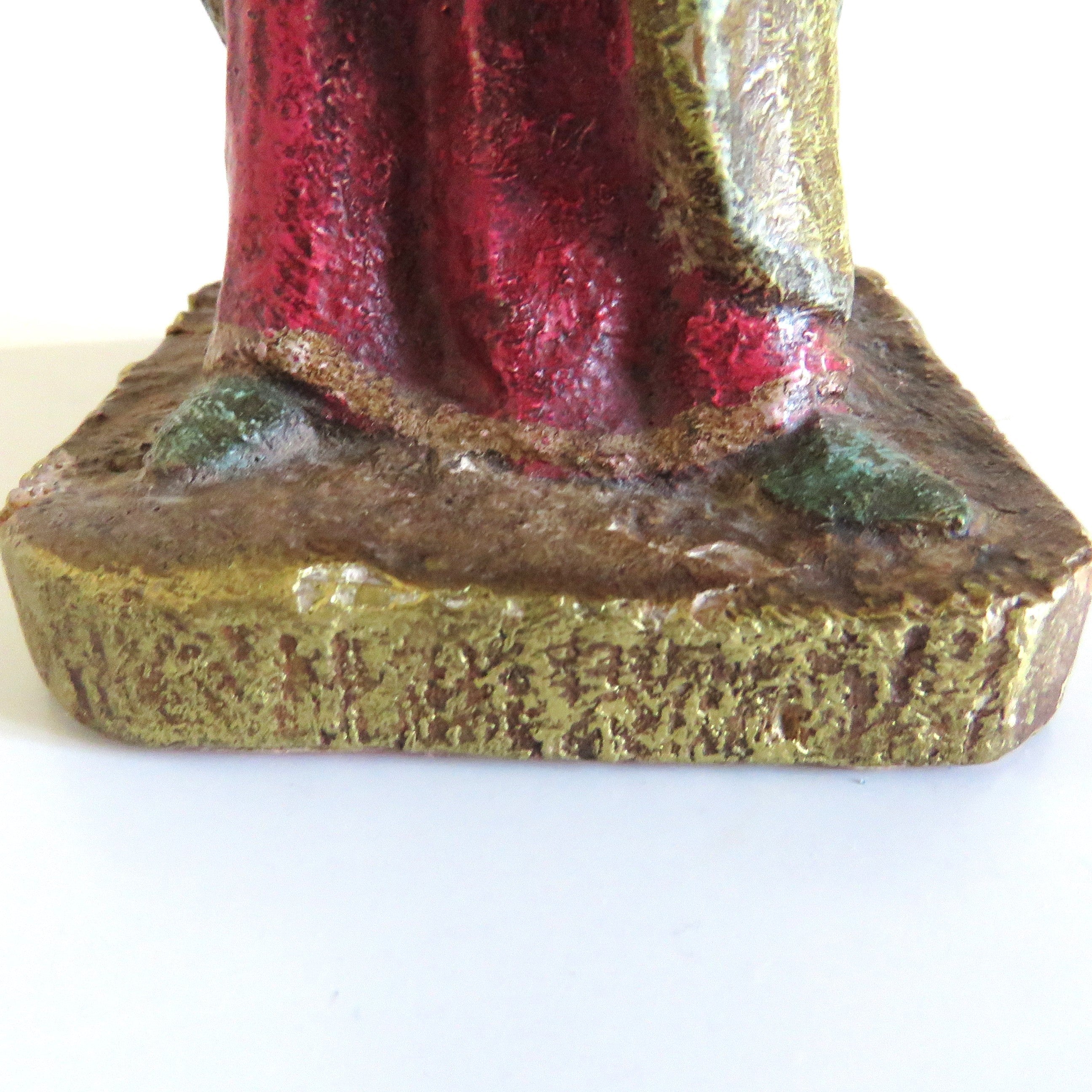 Close-up of the base and lower robe of a figurine, showing a textured gold base with a red and green garment, on a plain white background.