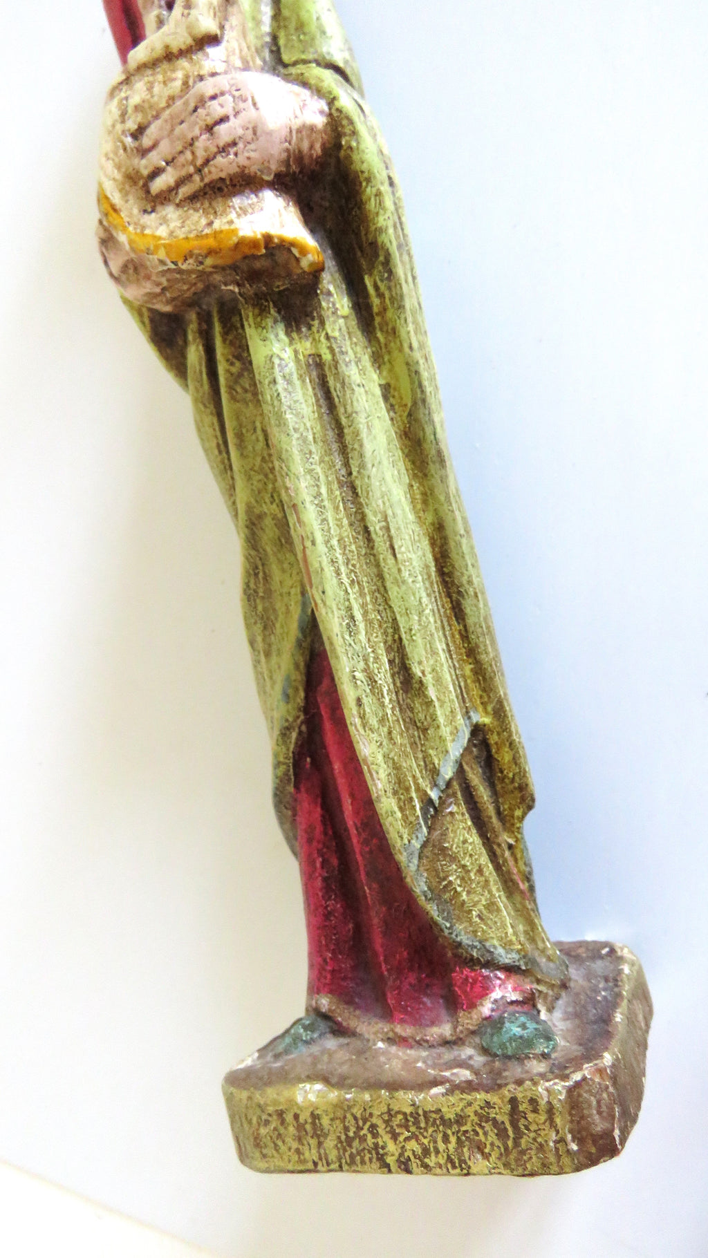 A close-up of a hand-painted wooden religious statue showing the lower half of a robed figure with green, red, and gold detailing, standing on a textured base against a plain white background.