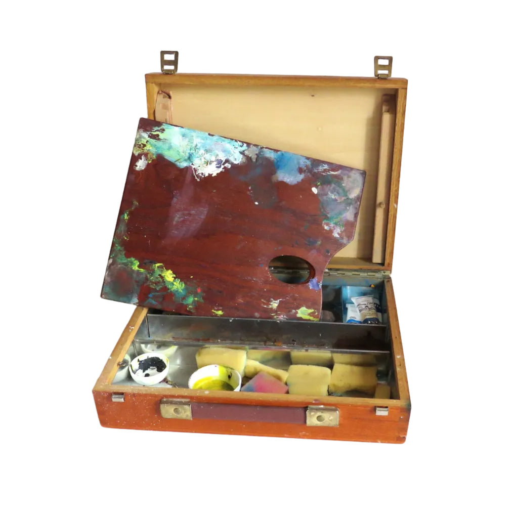 An open vintage wooden artist palette case on a white surface, with a large multicolored paint palette propped inside and small paint tubes visible in the compartment below.