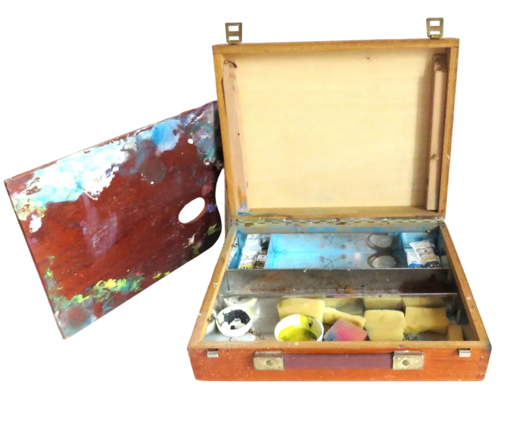An open wooden artist's palette case with brass hardware, showing paint wells, brushes, and a palette with multicolored paint, set against a plain white background.