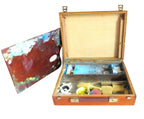 An open wooden artist's palette case with brass hardware, showing paint wells, brushes, and a palette with multicolored paint, set against a plain white background.