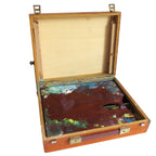 An open vintage wooden artist's palette case with brass latches, showing a stained multicolor paint palette inside on a white floor against a white wall.