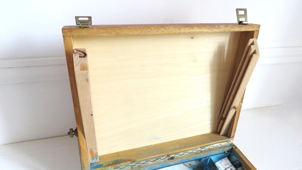 An open wooden storage box or case with a light natural wood interior and metal hinges, photographed against a plain white wall and floor.