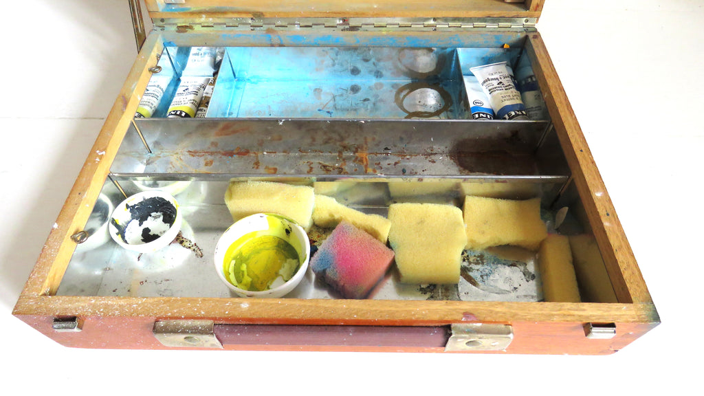 An open wooden artist's palette or storage case with metal trays, showing paint tubes, brushes, yellow sponges, a small cup of yellow liquid, and other small items on a white background.