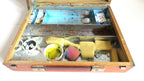 An open wooden artist's palette or storage case with metal trays, showing paint tubes, brushes, yellow sponges, a small cup of yellow liquid, and other small items on a white background.