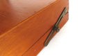 Close-up of a warm brown wooden box corner with a dark metal latch and screw closure, photographed against a plain white background.