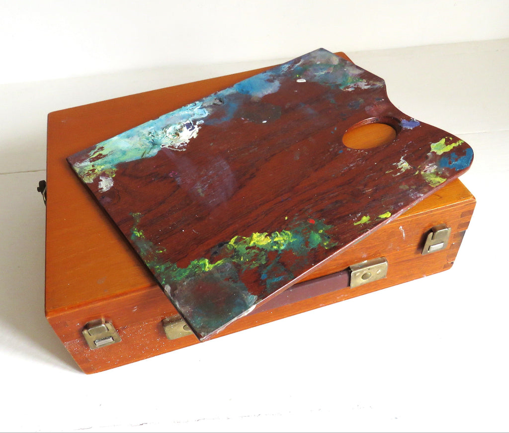 A vintage wooden artist's palette resting on top of a matching wooden case, both showing signs of use with multicolored paint splatters, set against a plain white background.