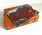 A vintage wooden artist's palette resting on top of a matching wooden case, both showing signs of use with multicolored paint splatters, set against a plain white background.