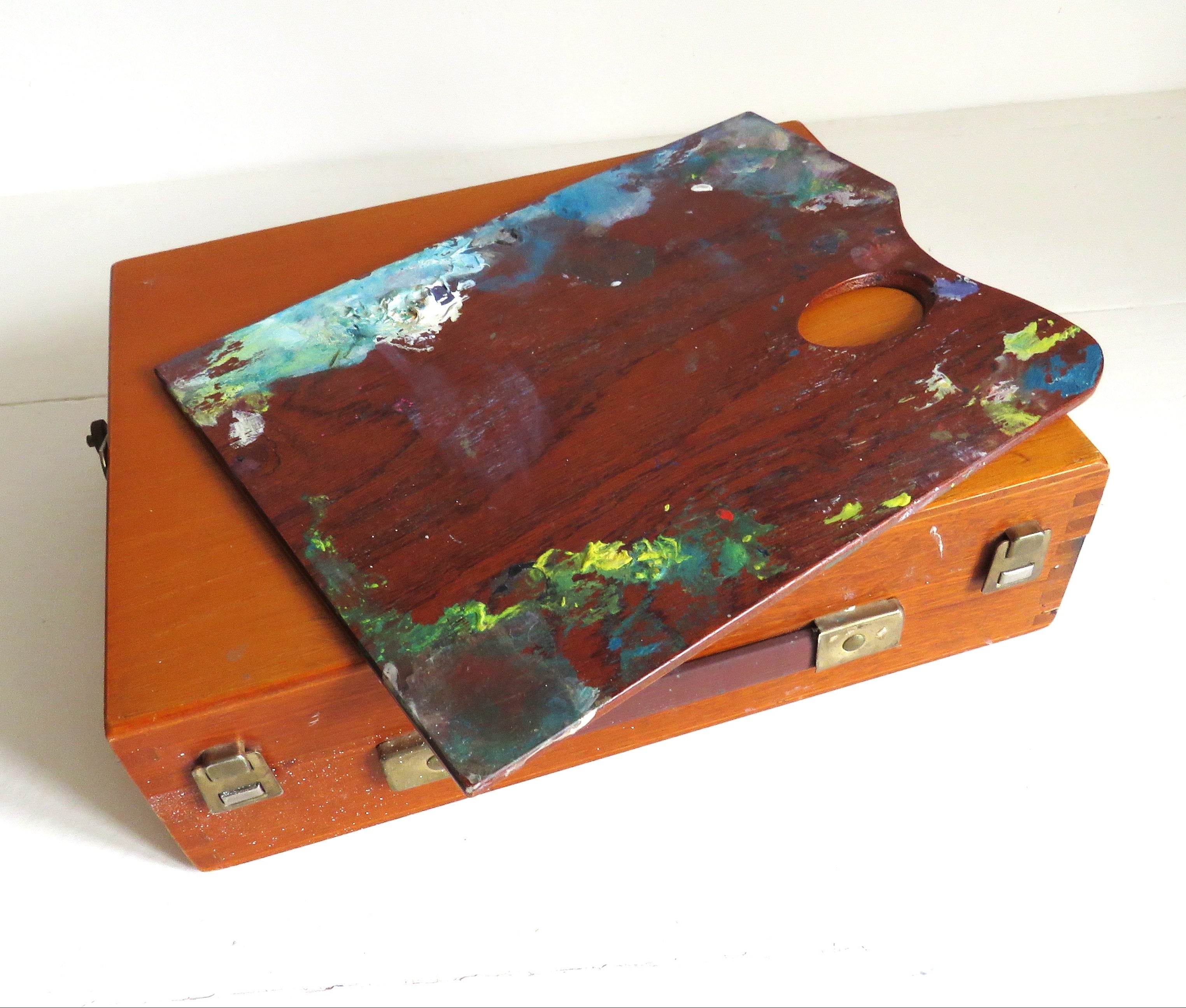 A vintage wooden artist's palette resting on top of a matching wooden case, both showing signs of use with multicolored paint splatters, set against a plain white background.