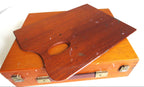 A wooden artist palette with a cutout handle resting on top of a matching wooden case with brass latches, photographed against a plain white background.
