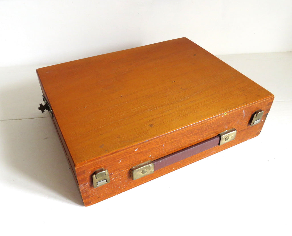 A vintage rectangular wooden art box with a warm orange-brown finish, brass-toned metal latches, and a dark handle, photographed on a plain white surface.x