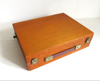 A vintage rectangular wooden art box with a warm orange-brown finish, brass-toned metal latches, and a dark handle, photographed on a plain white surface.x