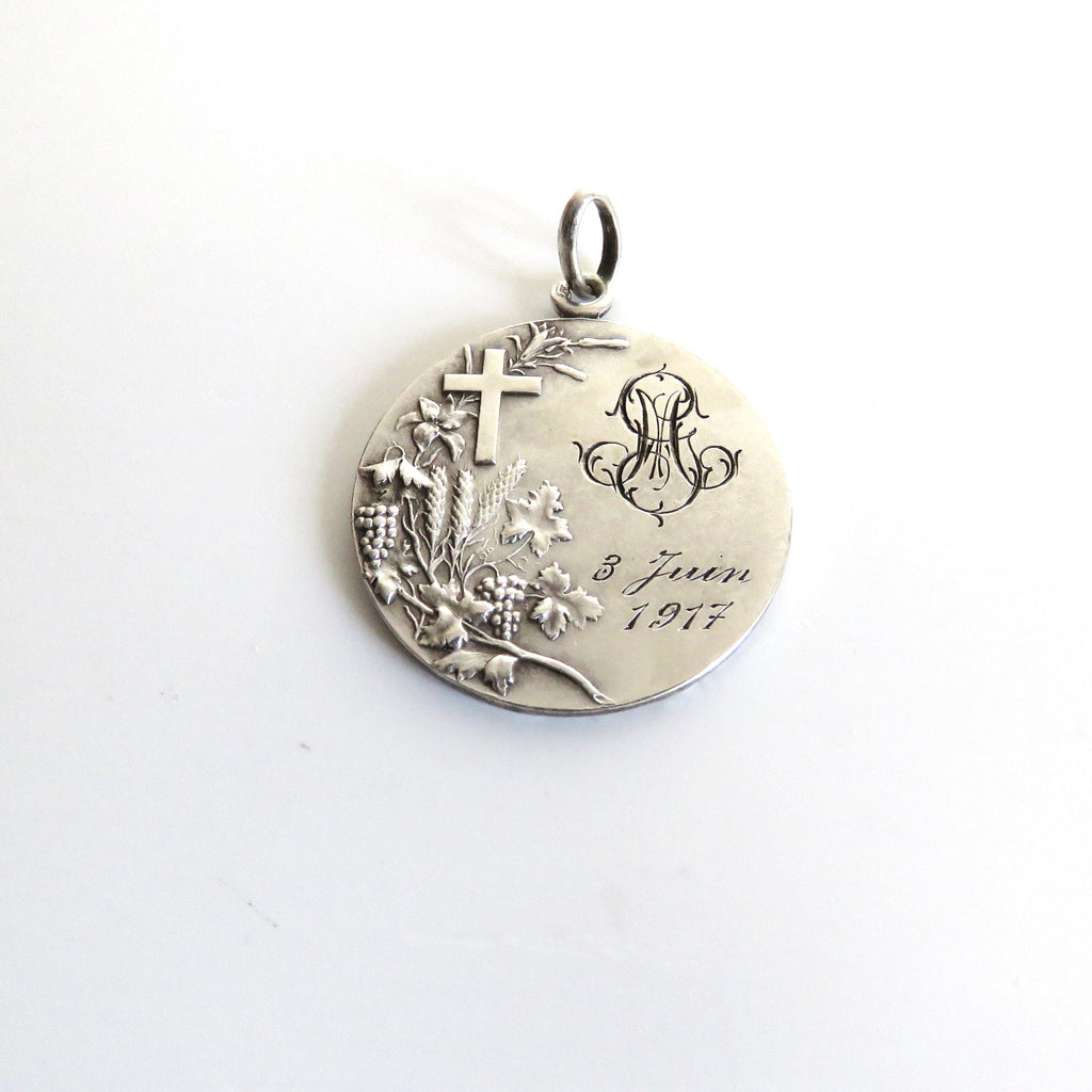 A round silver pendant with an engraved cross, floral motif, monogram, and the text '9 Juin 1917', shown on a plain white background.