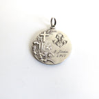 A round silver pendant with an engraved cross, floral motif, monogram, and the text '9 Juin 1917', shown on a plain white background.