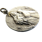 A round silver pendant featuring a raised relief carving of a kneeling figure, photographed on a smooth gray surface.