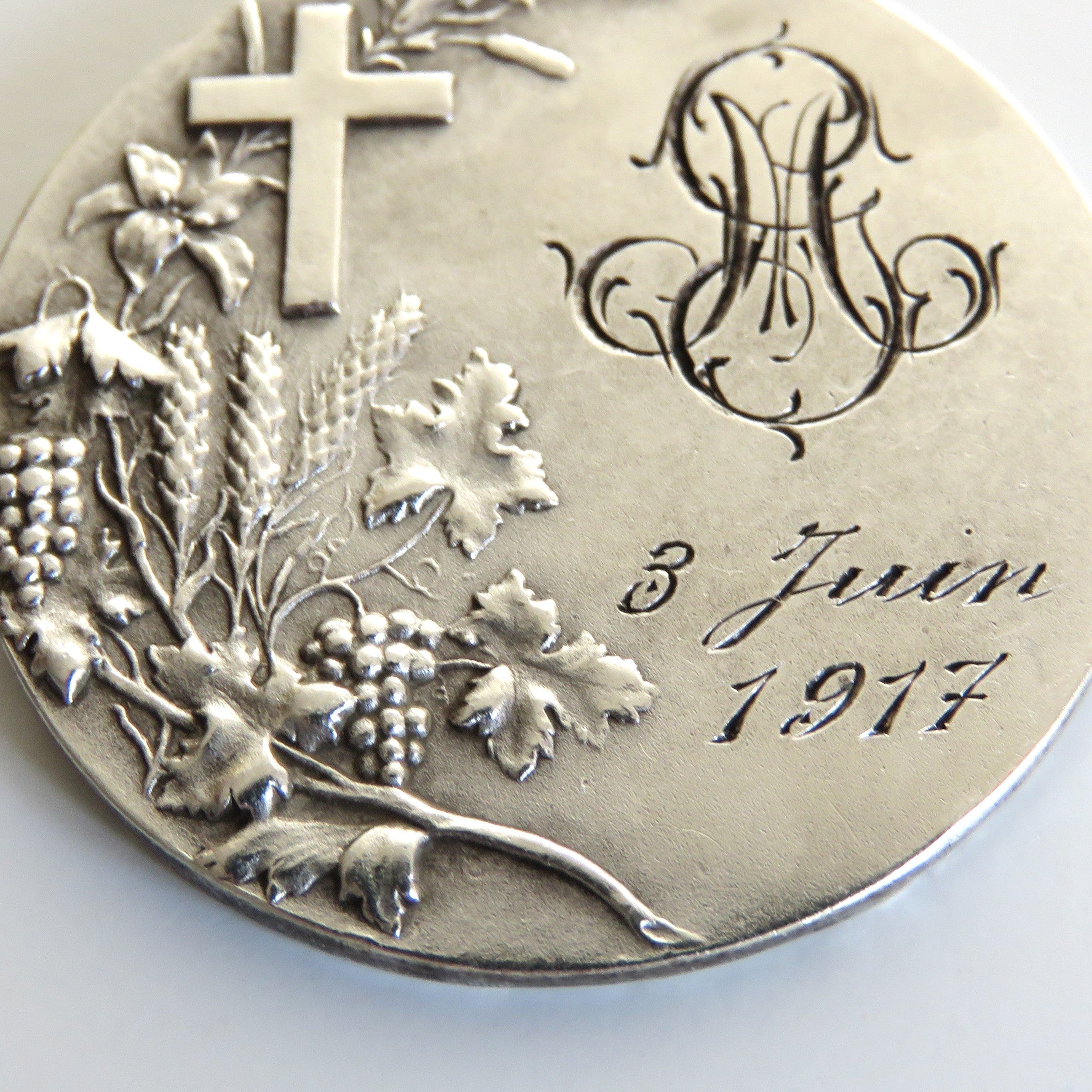 A round silver commemorative coin or medallion with an embossed cross, grapevine leaves and grapes, a monogram, and the inscription '3 Juin 1917' on a plain light background.