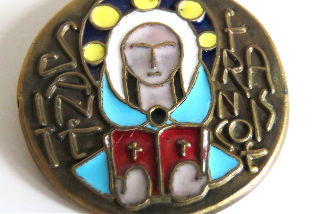 A round vintage religious medallion featuring a stylized depiction of the Saint Francois with gold-tone metal, light blue enamel, red enamel, and yellow enamel details on a white background.
