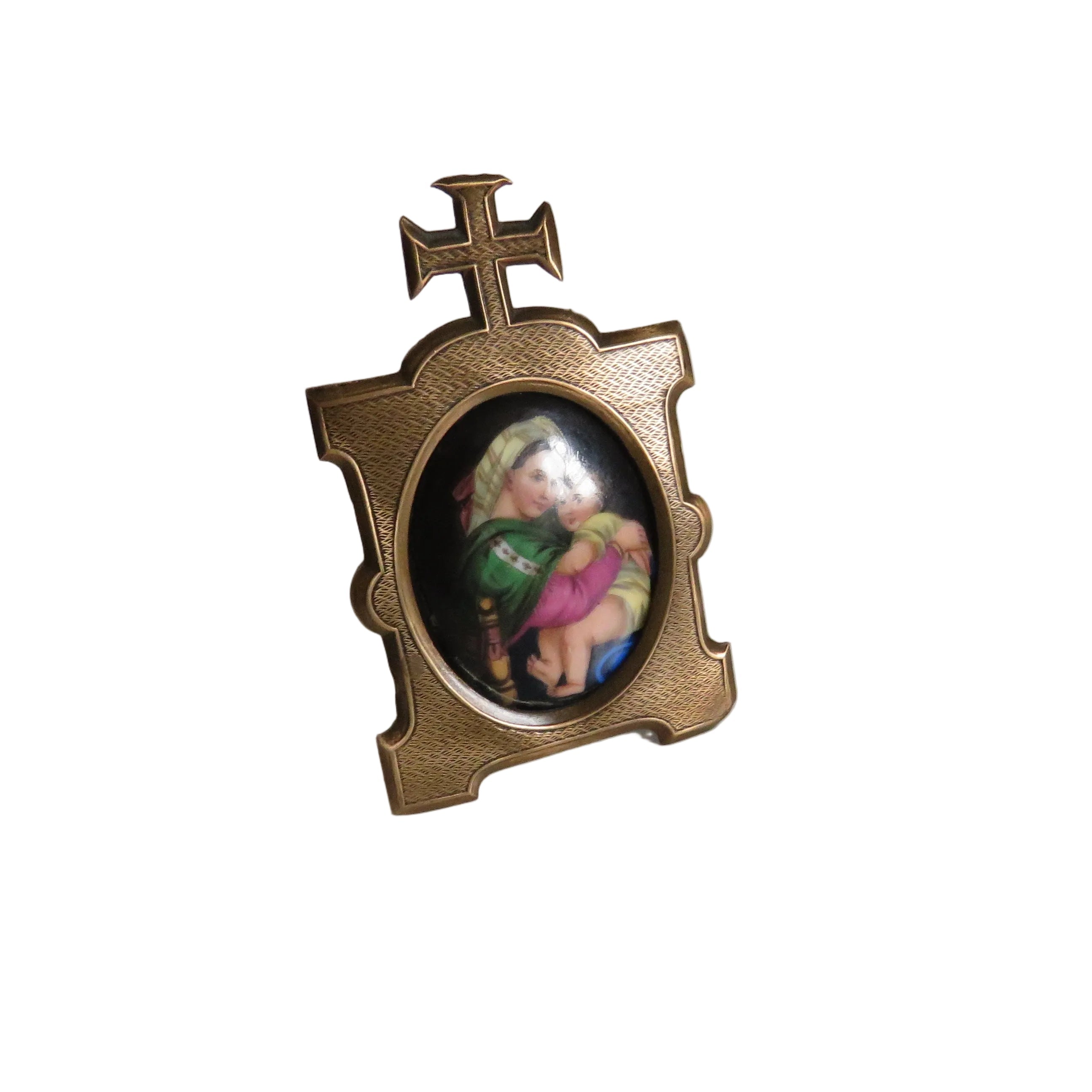 A small decorative tabletop cross with an oval inset containing a colorful religious illustration, photographed on a plain light gray surface against a white wall.