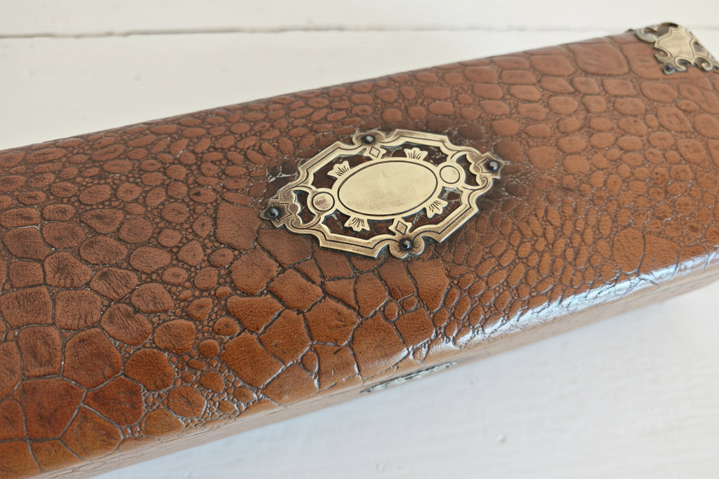 A close-up of a brown textured leather or faux-croc case with an ornate antique brass oval clasp on a white background.