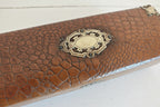 A close-up of a brown textured leather or faux-croc case with an ornate antique brass oval clasp on a white background.
