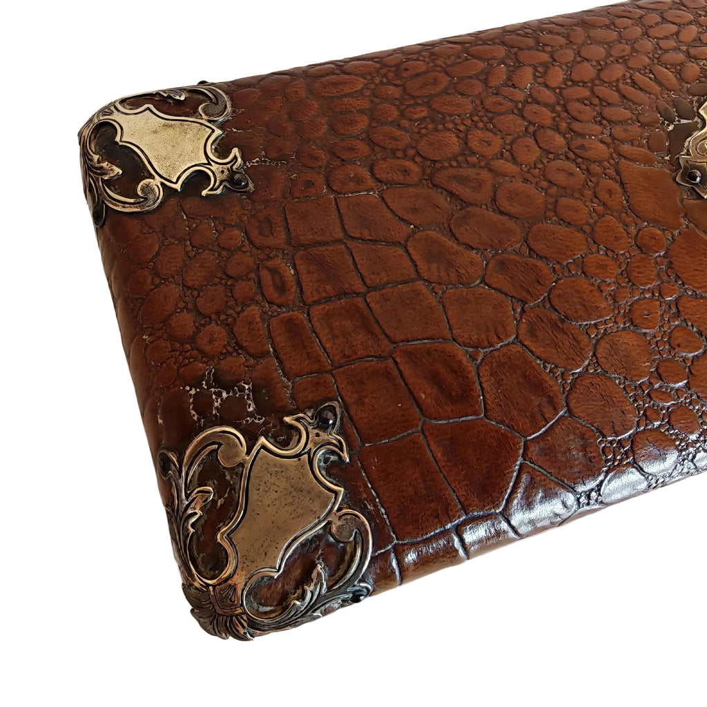 A close-up of a brown crocodile-embossed leather box or with decorative antique bronze corner protectors, resting on a plain off-white surface.